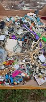 Wide overhead photo of a large box filled with assorted costume jewelry including necklaces, bracelets, and pins in a mixed pile.