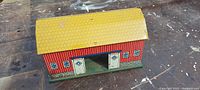 Front and side view of small antique tin toy barn on a wooden surface, showing yellow roof and red walls with door and window patterns