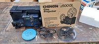 Chinon 2500GL cine projector, power cord, two metal reels, movie canister, and original cardboard box on wooden table.