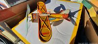 Top down angled view of orange and yellow tin toy airplane sitting on a white surface with yellow border, showing wings and tail detail.
