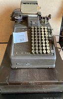 Front view of a dark metal antique adding machine showing the Burroughs brand on the front and the machine positioned on a wood base.