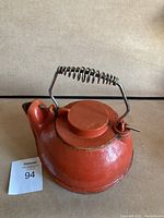 Red cast iron kettle with circular lid and metal spring handle, sitting on a table with a lot number card.