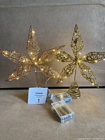 Two gold star-shaped Christmas tree toppers with battery operated lights, one wired gold, the other gold sparkles, displayed standing with visible batteries and wiring.