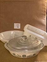 Three pieces of Glassbake bakeware arranged on a surface, showing the Green Daisy floral pattern, including one oval covered bake dish, one loaf pan, and one divided oval dish.