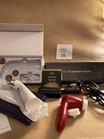 White suction beauty device, NYX matte bronzer, black 3-in-1 straightener brush, red face scrubber with interchangeable heads, blue wallet laid out on a beige surface.