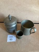 Photo showing pewter tobacco container, empty pewter mug, and pewter shot glass together on a surface with a lot number card