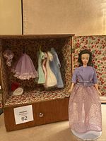 Full view of Mitzi doll standing inside wooden homemade closet showing floral fabric lining and multiple hanging handmade outfits