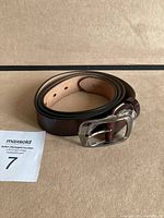 Rolled-up dark brown genuine leather women's belt with a shiny silver buckle, photographed on beige background.