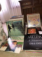 Multiple vinyl LP albums stacked in wire holder and on floor; visible front covers include Simon & Garfunkel, Peter, Paul & Mary, Mitch Miller Christmas album, Glenn Miller album; shows condition and selection of records in lot.
