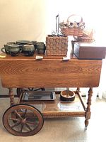 Front and top view showing the solid oak wood grain, folded side leaves, large wagon wheel style wheels, lower shelf with glass trays, and items on top not included in sale.