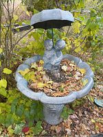 Front angled view of the bird bath showing concrete basin with scallop edge and metal figurine of children holding umbrella at center, leaves inside basin