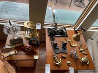 Photo showing small table lamp, multiple clocks on tables by window, and brass decor items spread out on another table.