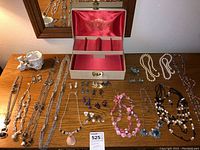 Full overview of jewelry collection on wooden surface including necklaces, earrings, and jewelry box with pink silk lining.