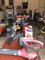 Overall view of Craftsman miter saw, DeWalt reciprocating saw, Dirt Devil Hand Vac box, red leaf blower with cords on the table and other items around such as paint brushes and golf clubs.