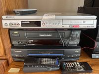 Front view of Pioneer DVD recorder DVR-219 (silver), JVC VHS player HR-S7600U (black), and Panasonic VHS player NV-HS960 (black) stacked with cables and remotes in front.
