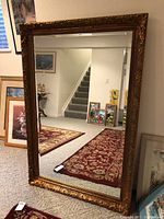 Full view of large rectangular gilded mirror leaning against a wall showing the mirror's size and appearance in a room with carpet and framed photographs nearby.