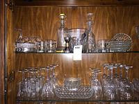 Two shelves inside wood cabinet holding various glass and crystal items including decanters, stemware glasses, pitcher, serving plates and bowls.