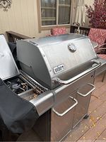 Full view of Weber Genesis stainless steel natural gas grill on patio with red chairs visible behind