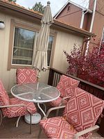 View of full patio set including round glass table, umbrella, and four chairs with red patterned cushions outdoors on wood deck beside house.