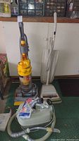 Three vacuums standing next to a cabinet: upright Dyson with yellow dustbin, vintage beige Eureka upright, and mint green Kenmore canister vacuum with hose.