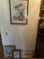 Three framed prints: floral prints and a print with bird and flowers, arranged on floor with wood wall behind.
