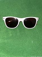 Front view of white framed sunglasses with dark lenses on green background, showing frame shape and lens condition.