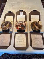 The three Norman Rockwell collector plates with certificates of authenticity and booklets in original boxes displayed on a table.
