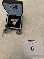 Three sterling silver brooches displayed on a light tablecloth; one in Bond Boyd box is a trillium flower, the others are roses cluster and a scotch thistle with amber