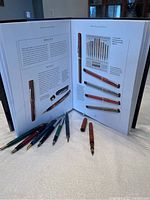 Photo showing seven collectible antique fountain pens placed on table in front of open collector's guide book with detailed vintage pen images and text.