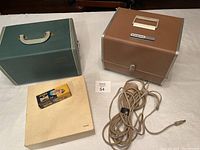 Argus 500 Electromatic slide projector in brown textured carrying case, Kodak Carousel Slide Tray box, green case for slide changer magazines, extra lamp box on white surface