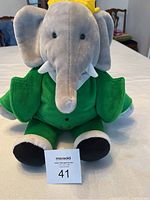 Front view of the grey elephant plush wearing green jacket and yellow crown, seated on white surface.