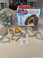 Container filled with various metal cookie cutters and a plastic round cookie mold alongside the Betty Crocker Baken Fill Bake Set box on a table.