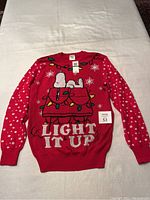Full front view of the red Peanuts Christmas sweater with Snoopy lying on decorated doghouse, white snowflakes and 'LIGHT IT UP' text.
