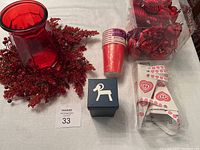Photo showing a red glass vase surrounded by a red wreath of berries and foliage, stacked red disposable cups, a blue gift box, and a clear box of red ornaments.