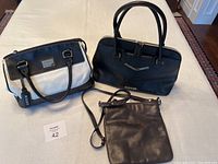 Three purses displayed together on wooden floor background: white/black Tignanello purse, black DKNY handbag, black Kenneth Cole crossbody purse