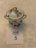 Jam jar with cherry design and gold trim, placed on a cream patterned surface with auction lot number card visible.
