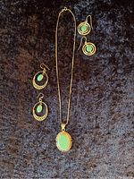 Top-down image showing two pairs of earrings and one pendant necklace with green stones, arrayed on dark fabric.