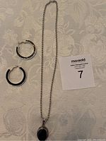 Sterling silver chain necklace with black gemstone pendant and two sterling silver hoop earrings laid flat on a white patterned tablecloth.
