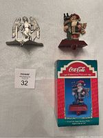 Photo showing both stocking holders with Coca-Cola Santa in original box and pewter angel holder side by side on surface.