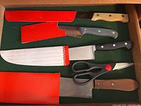 Top-down view of four knives and a pair of scissors in a drawer, each knife with protective red blade sheaths, showing handles and blade shapes.