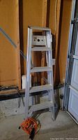 5-foot aluminum ladder standing against a wooden wall, orange extension cord and work light coiled at its base.