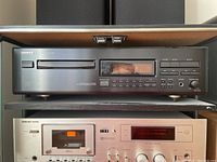 Front view of Onkyo DX-7210 CD player showing horizontal disc tray, digital display with orange lighting, track control buttons, and power switch on left side. Black metal panel with labeling.