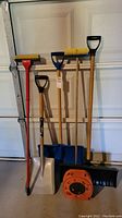 Full view of four snow shovels, mop, cleaning brush and orange extension cord against garage door