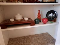Shelf with Asian style white ceramic serving set on wooden tray, two stone apples (green and red), orange double-gourd vase on black stand, and black decorative plate on stand.