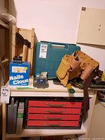 Overview of tools on white shelf including axe, tool belt, drill case, box of nails, smaller hand tools on top of red metal storage chest.