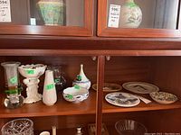 Glass and ceramic vases alongside ceramic decorative plates and bowls in wooden cabinet.