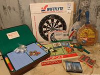 Overview of the lot showing Swiftflyte dart board box, Monopoly games partially visible, vintage Mah Jong set open with tiles and dice, Rubik's Cube, slingshot, Rallyball paddles, and cards.