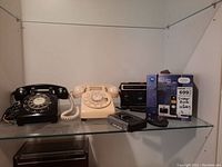Shelf with two vintage rotary dial telephones (black and cream), Realistic radio and cassette player, Sony earpods, Aiwa cassette recorder, and Motorola DECT 6.0 cordless phone in box