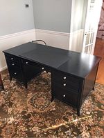 Front and side angled view of black Arhaus desk on patterned rug showing drawers and desk matt on top