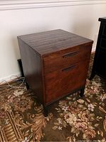 Front and side views of the Arhaus wood filing cabinet with metal base, showing the cabinet's wood grain finish, metal handles, two drawers, and visible staining on top surface.